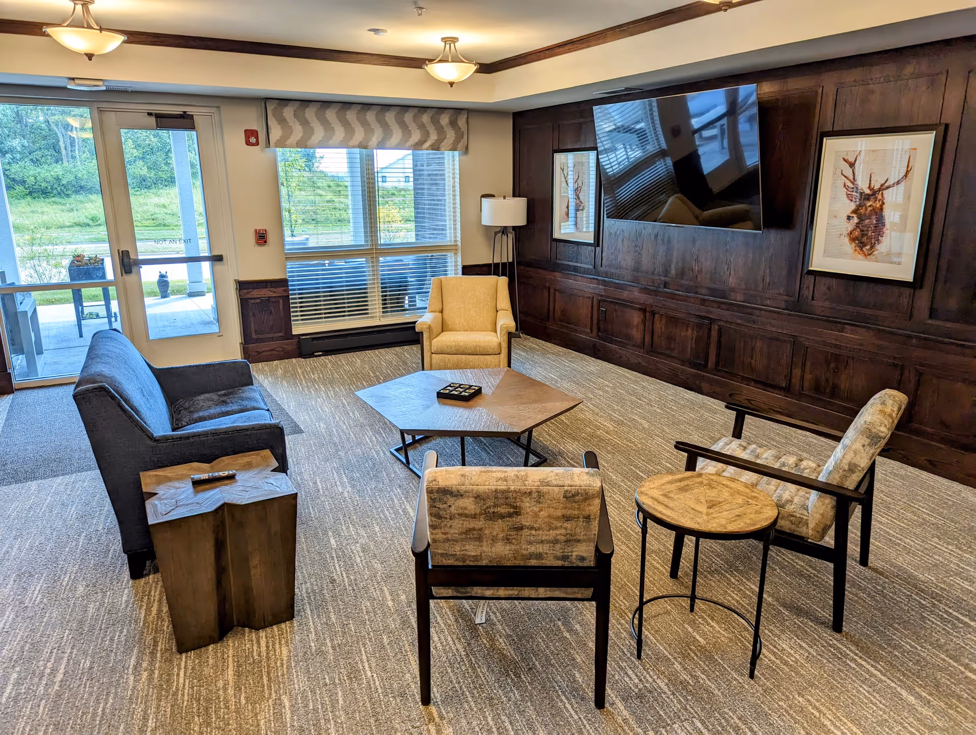 A cozy living room area with a dark gray sofa, two patterned armchairs, and a beige armchair arranged around a hexagonal wooden coffee table. There is a small round side table and a wooden side table with a remote control on it. The room has a large window with blinds and a glass door leading outside. The wall opposite the seating area features dark wood paneling, a mounted flat-screen TV, and two framed pictures of deer.