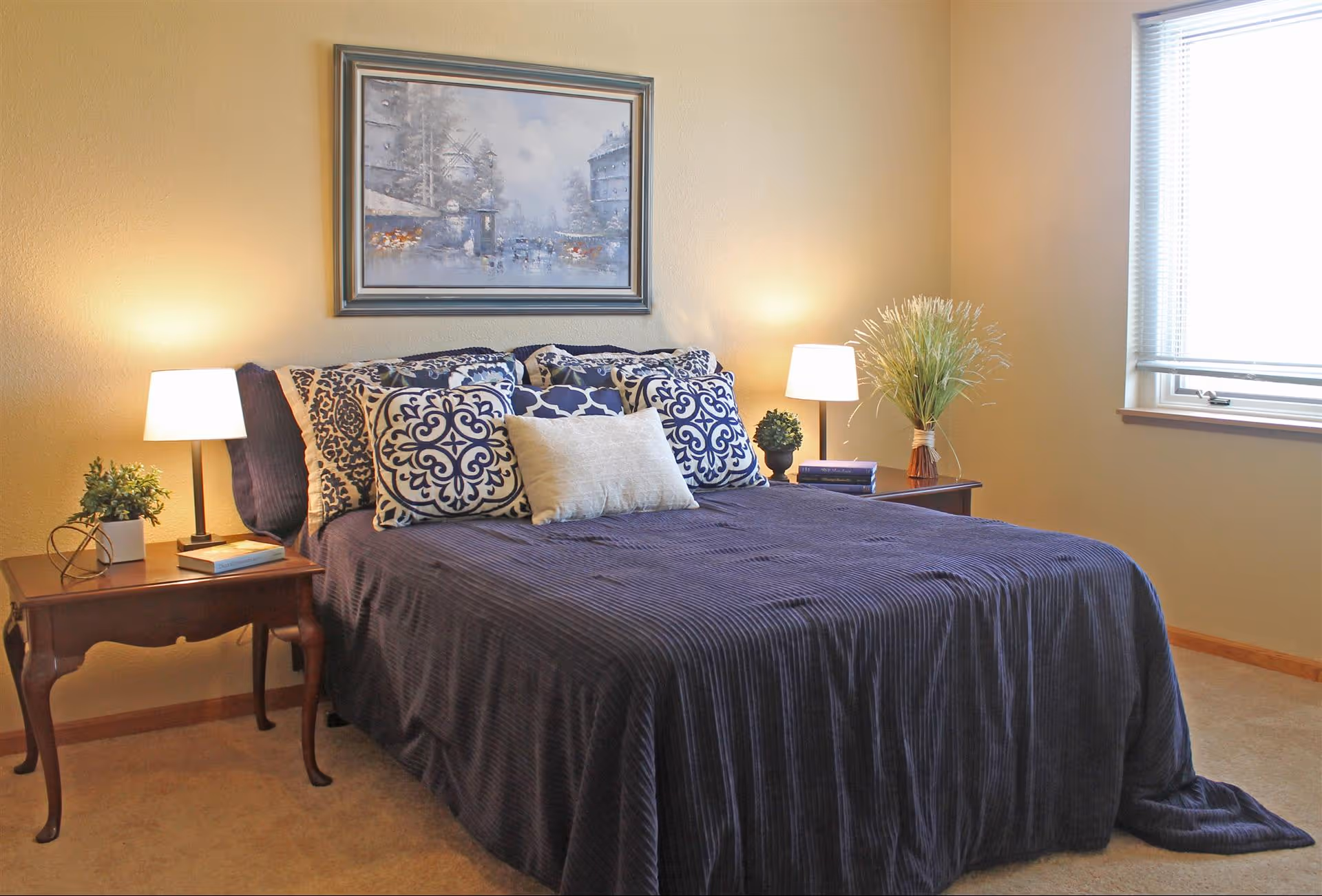 A neatly made bedroom with a navy bedspread and patterned pillows flanked by two bedside tables with lamps, plants, and a framed painting above the bed.