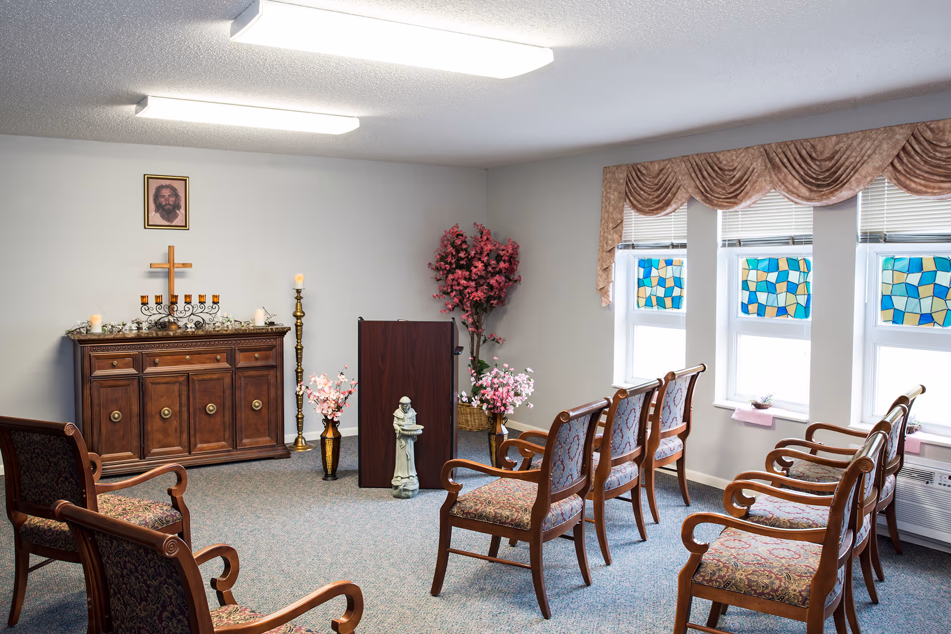 A small chapel room with wooden chairs arranged in rows facing a wooden podium. The room has a cabinet with a cross and candles on it, a framed picture of Jesus on the wall, and decorative flowers. Three windows with stained glass patterns and valances allow natural light into the room.