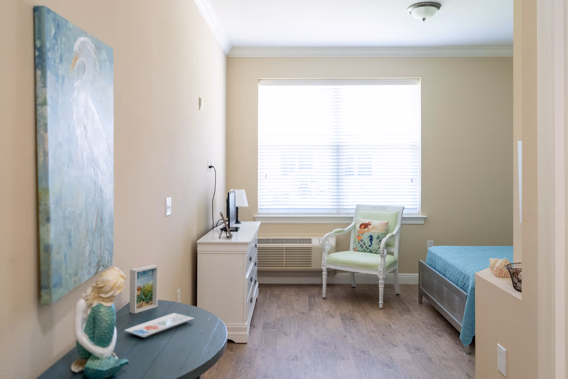 A bright and cozy bedroom with a large window covered by white blinds. The room features a white dresser with a small TV and lamp on top, a light green armchair with a decorative pillow, and a bed with a blue quilt. There is a round table with a mermaid figurine and framed artwork, and a large painting of a white bird on the wall.