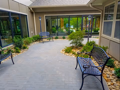 Small paved courtyard with metal benches, lamp posts, landscaping and large windows of the senior living facility.