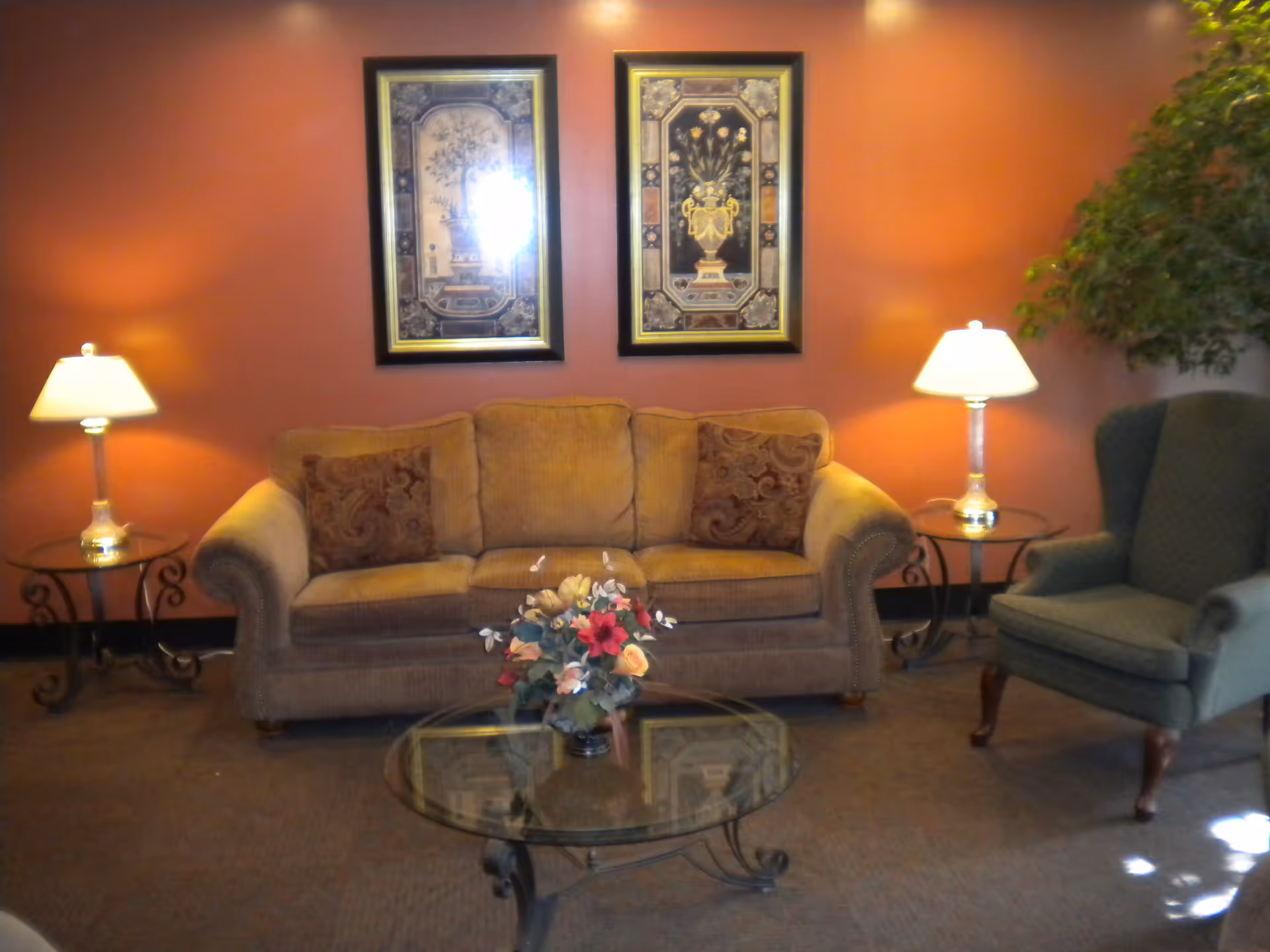 A cozy living room area with a brown couch adorned with two patterned pillows, a glass coffee table with a floral arrangement, two side tables each with a lamp, a green upholstered armchair, and two framed floral artworks on a warm orange wall.