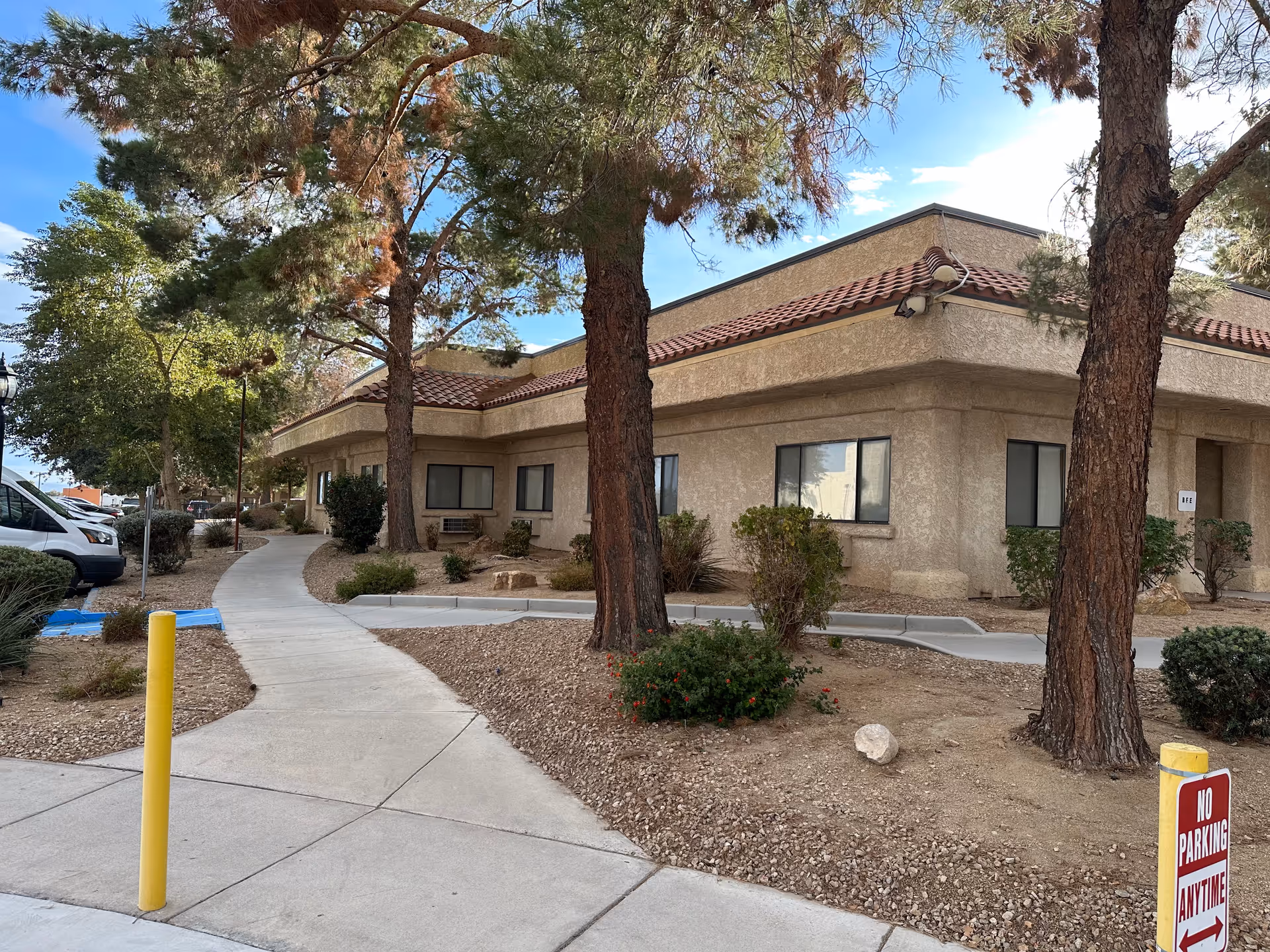 Single-story stucco nursing facility building with a tiled roof, sidewalk, trees, and desert landscaping at the entrance.