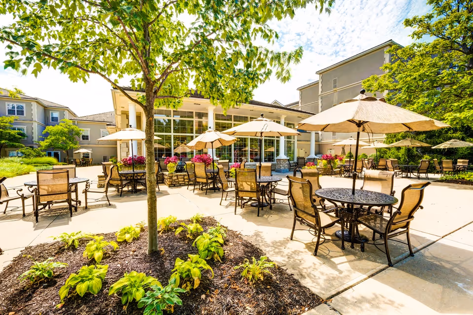 Sunny outdoor courtyard with metal patio tables, chairs, umbrellas, and landscaping in front of a senior living building.
