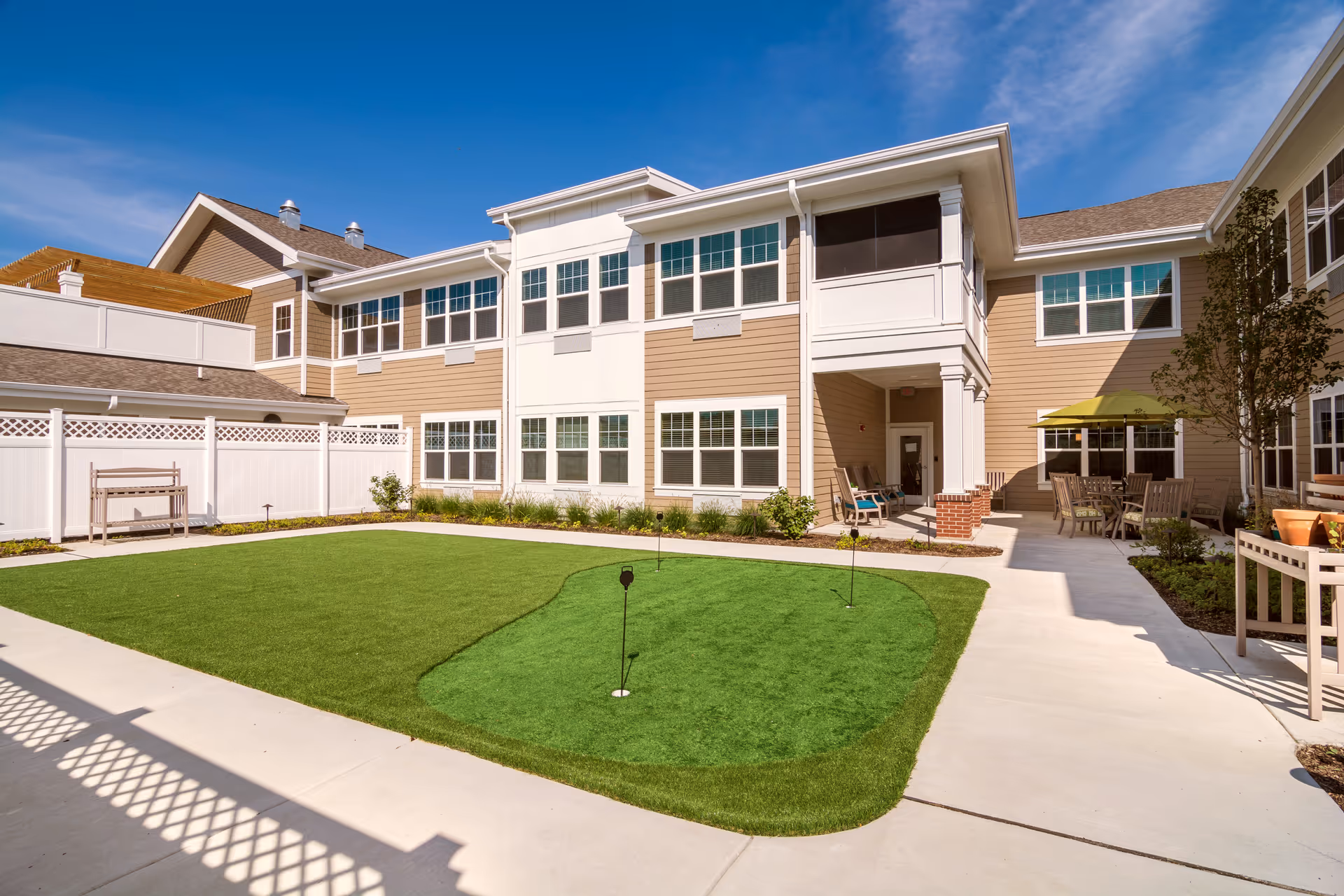 Outdoor courtyard area of a senior living facility with a small putting green, surrounded by a beige two-story building with many windows. There are chairs and tables with umbrellas on the right side and a white fence on the left side under a clear blue sky.