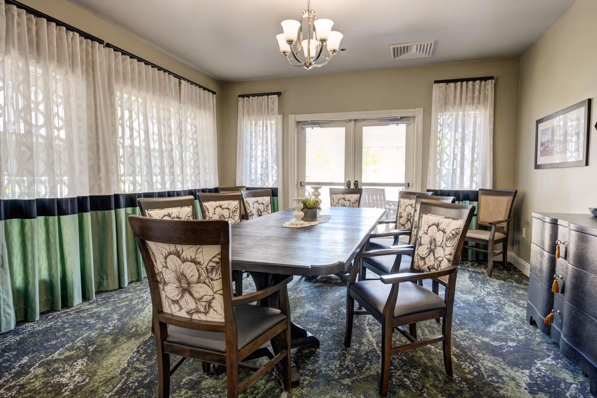 A dining room with a long rectangular wooden table surrounded by eight wooden chairs with floral patterned cushions. The room has large windows with sheer white and green curtains, a chandelier hanging from the ceiling, and a sideboard with decorative items on the right side. The floor is carpeted with a green and blue patterned carpet.
