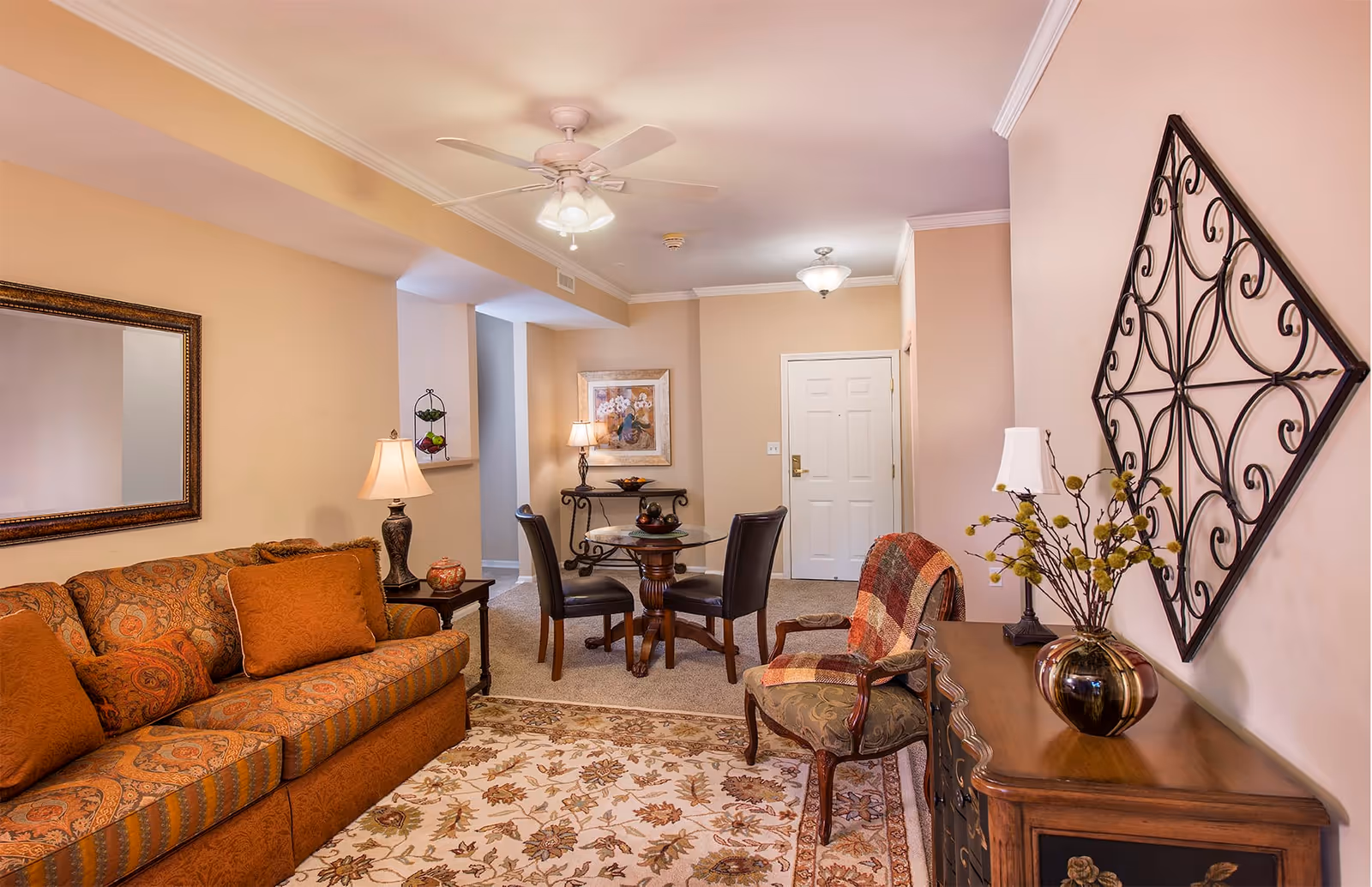 A cozy living room area with a patterned orange sofa adorned with matching cushions, a wooden side table with a lamp, and a decorative mirror on the wall. There is a round glass-top dining table with four chairs in the background near the entrance door. An upholstered armchair with a plaid throw blanket is positioned next to a wooden cabinet with a vase of flowers and a table lamp. The walls are painted beige, and the room is well-lit with ceiling lights and a ceiling fan.