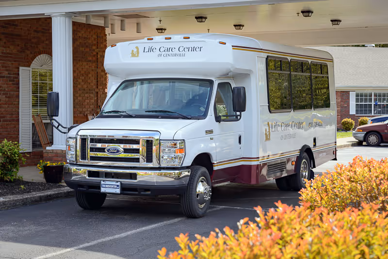 A white shuttle bus parked in front of a brick building with white columns. The bus has the logo and name 'Life Care Center of Centerville' on the front and side. There are bushes with orange and green leaves in the foreground.
