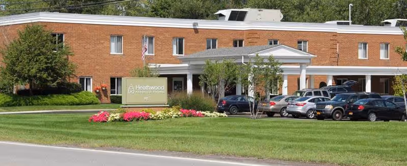 Exterior view of Heathwood Assisted Living and Memory Care facility, a two-story brick building with several windows, a white entrance canopy, a green lawn with flower beds, and a parking area with multiple cars.