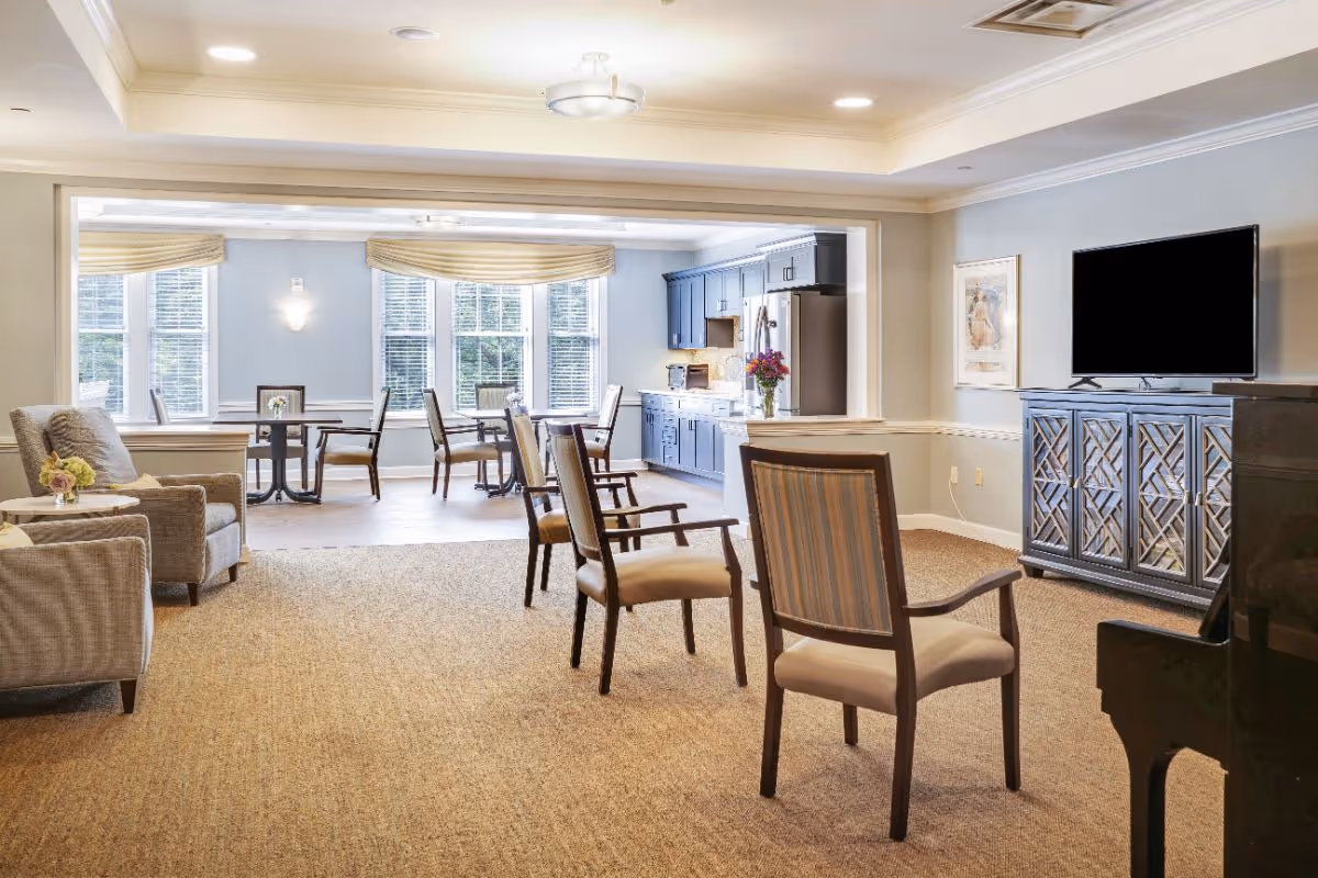 A bright and spacious common area in a senior living facility featuring a living room with several chairs arranged facing a flat-screen TV on a decorative cabinet. Adjacent to the living room is a dining area with tables and chairs near large windows with beige valances, and a kitchen area with blue cabinets and stainless steel appliances.