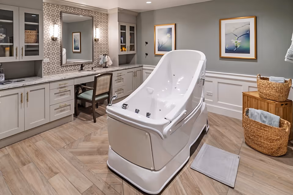 A modern bathing room featuring a large white therapeutic bath tub in the center, surrounded by light gray cabinetry with a marble countertop, a chair, and a large mirror on the wall. The room has wooden flooring, two framed floral artworks on the wall, and wicker baskets holding towels.