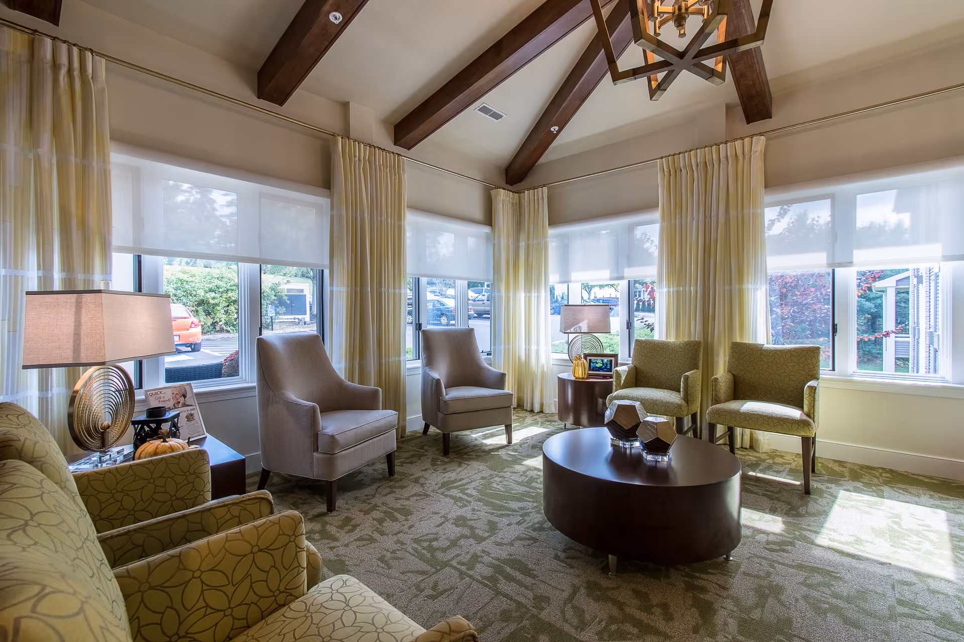 A sunlit communal sitting room with multiple upholstered armchairs around a central coffee table, large windows and exposed wooden ceiling beams.