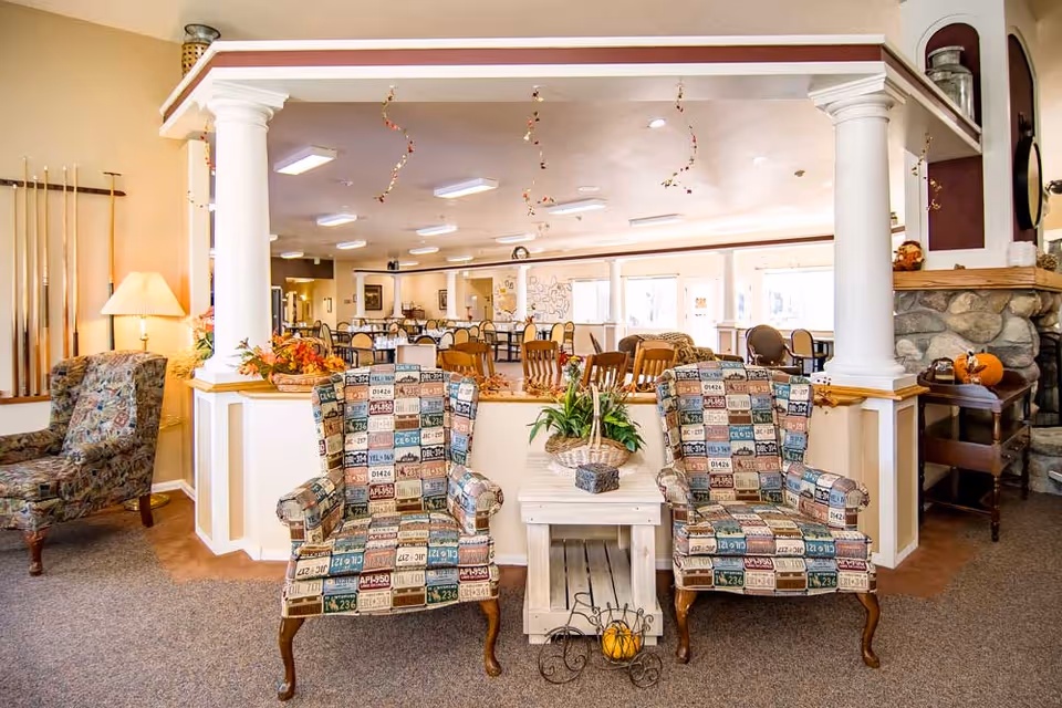 A cozy seating area with two patterned armchairs featuring a license plate design, separated by a small white wooden table with a basket of plants on top. The area is framed by white columns and overlooks a spacious dining room with multiple tables and chairs. The room is warmly lit with ceiling lights and decorated with autumn-themed accents, including pumpkins and fall foliage. A stone fireplace and a lamp with a beige shade are visible on the sides.