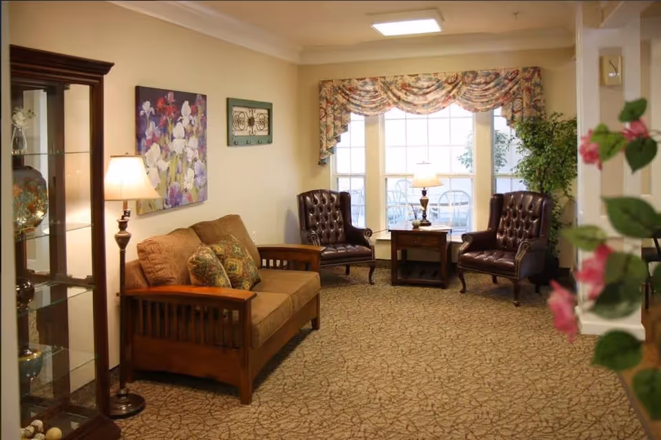 A cozy senior living facility lounge area with a brown cushioned wooden sofa, two dark brown leather armchairs, a wooden side table with a lamp, a glass display cabinet, floral wall art, and a large window with floral valance curtains. There are also some green plants and pink flowers partially visible in the foreground.