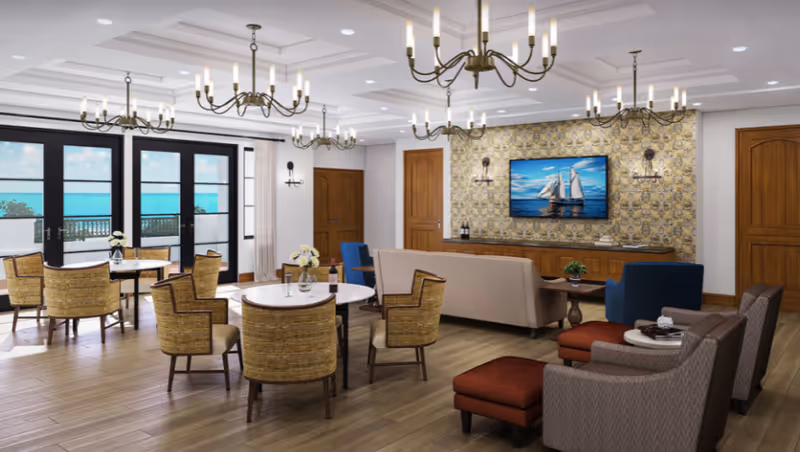 Spacious senior living common room with multiple seating areas, round dining tables, chandeliers, a TV on a patterned wall, and large doors opening to an ocean view.