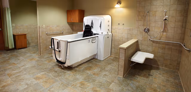 Accessible bathing room with an assisted lift bathtub, wall-mounted shower seat, grab bars, and tiled walls and floor.