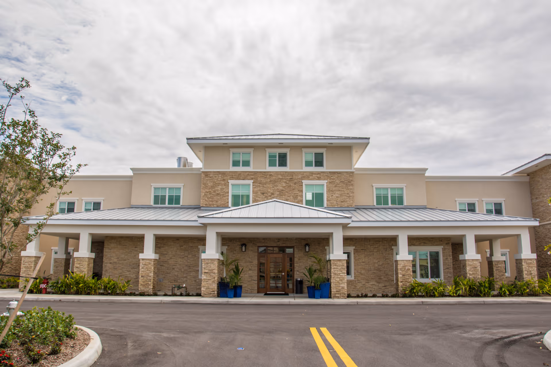Front exterior view of a modern senior living facility building with beige and stone facade, multiple windows, a covered entrance supported by columns, and landscaped greenery on either side of the entrance.