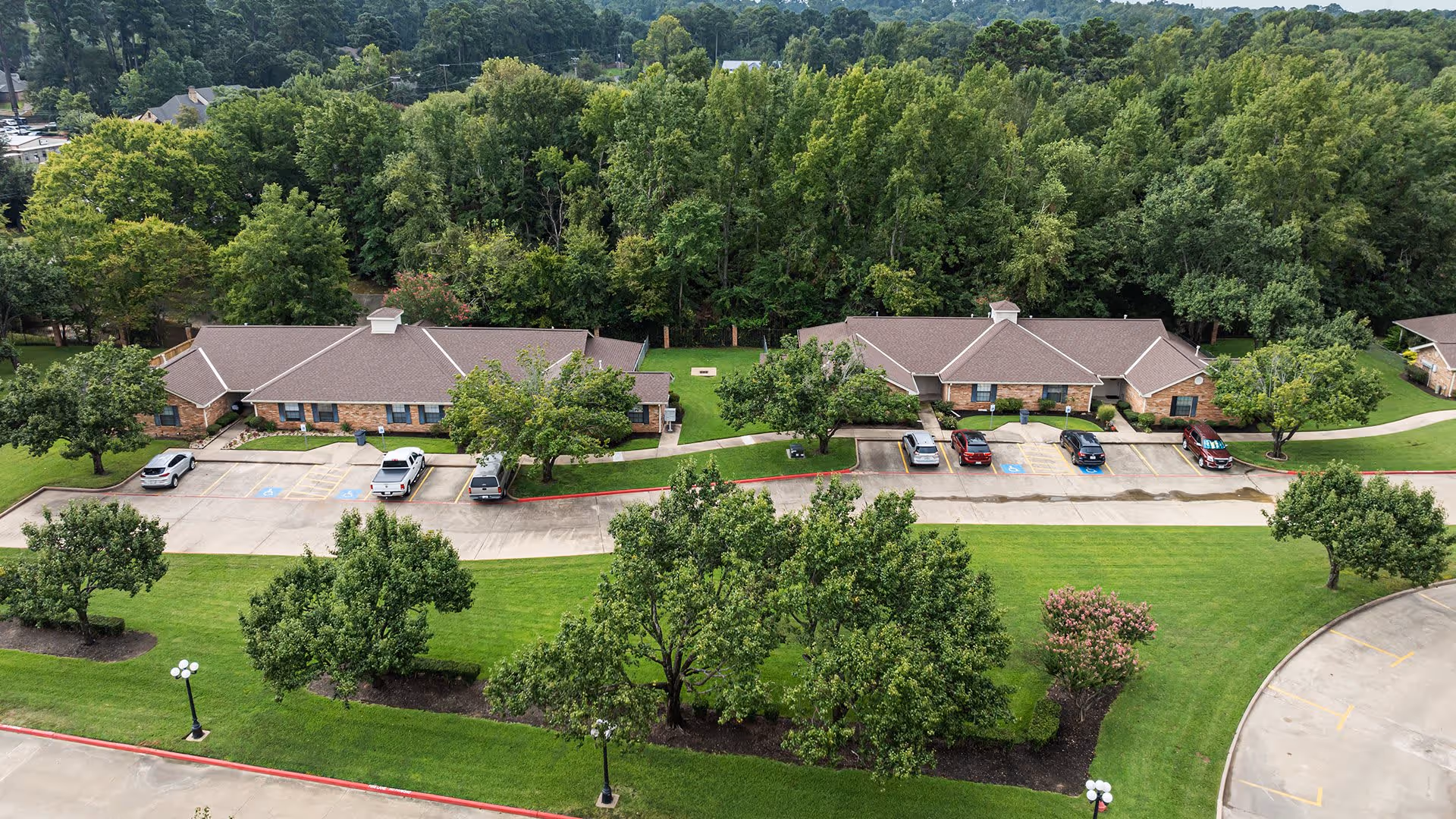 Aerial view of single-story brick senior living buildings with parking, landscaped lawns, and surrounding trees.