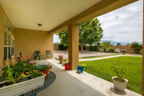 Covered patio area with several potted plants and two chairs, overlooking a green lawn, garden beds, and a tree with a brick wall and mountains in the background under a partly cloudy sky.