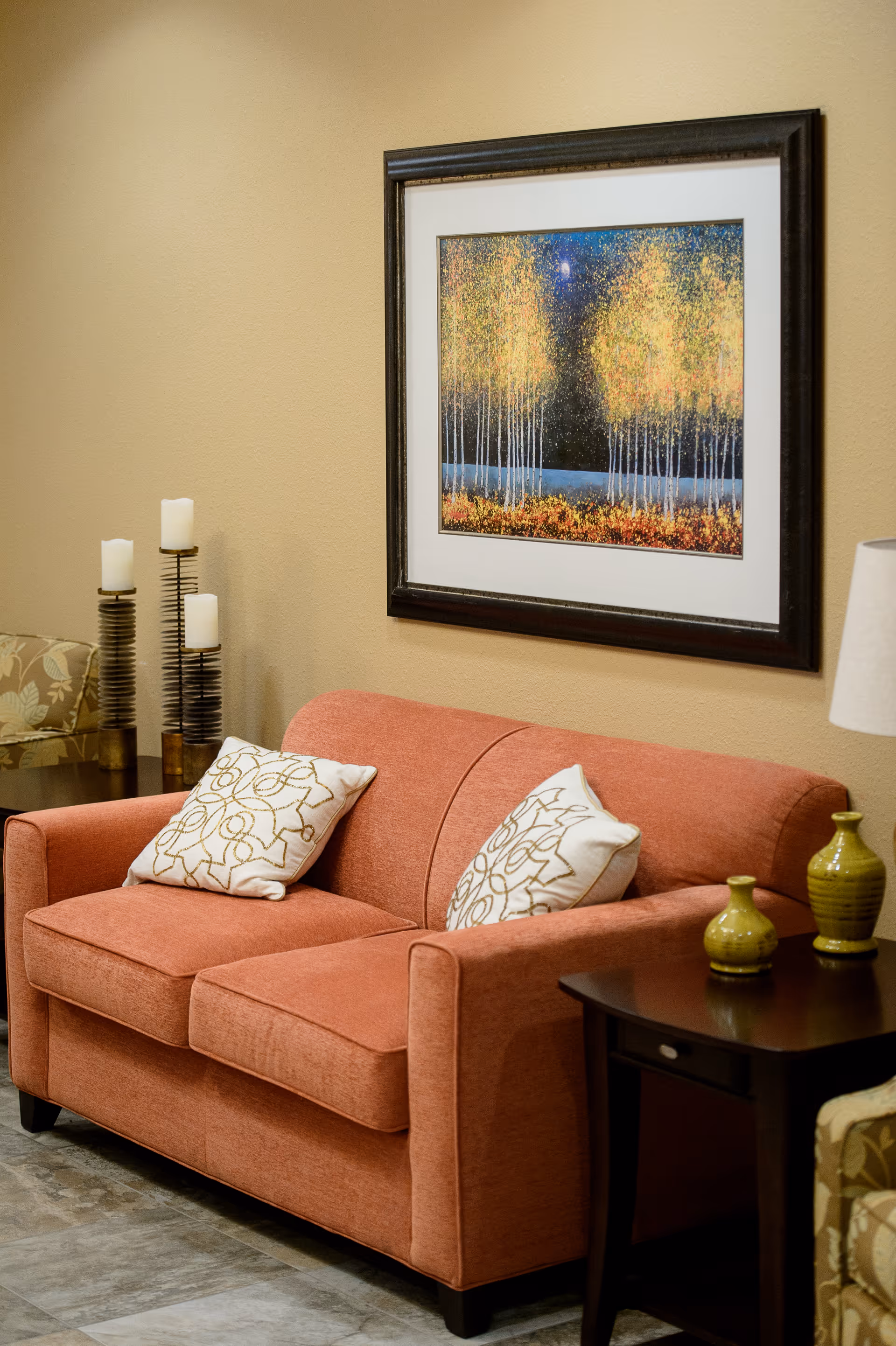 A cozy living room area with a coral-colored loveseat adorned with two white decorative pillows featuring gold patterns. Behind the loveseat is a beige wall with a framed painting of trees with yellow leaves against a dark sky. To the left, there are three tall candles on a stand and a floral-patterned chair partially visible. To the right, there is a dark wooden side table with two green decorative vases and a lamp with a white shade.