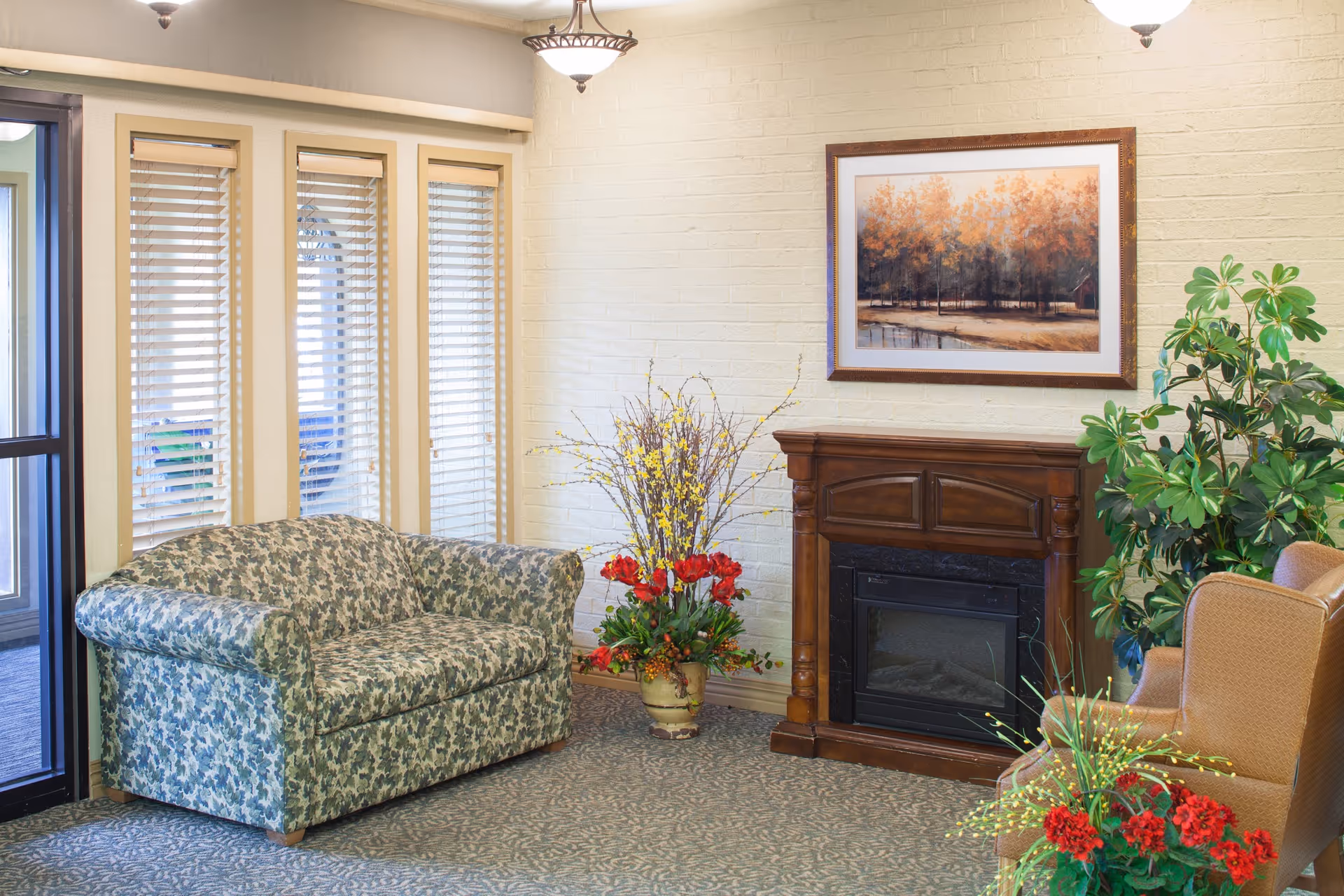 A cozy living room area with a floral-patterned loveseat, a wooden electric fireplace, and two beige armchairs. There are decorative plants and flowers around the fireplace, and a framed painting of autumn trees hangs above it. The room has a carpeted floor and windows with blinds letting in natural light.