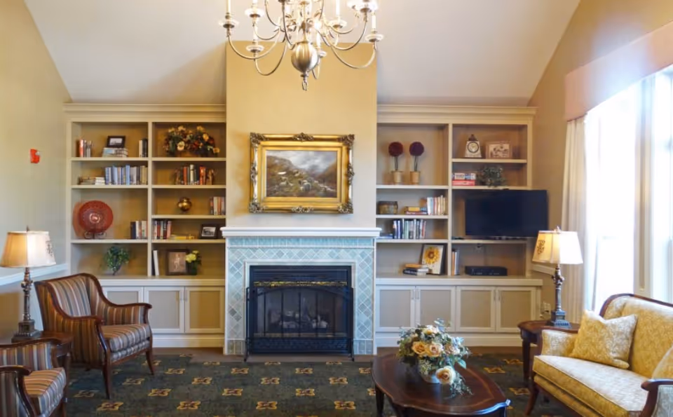 A cozy living room with a fireplace in the center, flanked by built-in shelves filled with books and decorative items. There are two striped armchairs on the left, a yellow patterned sofa on the right, a wooden coffee table with a floral arrangement in the middle, and two table lamps on either side. A chandelier hangs from the ceiling, and large windows with curtains allow natural light to enter the room.