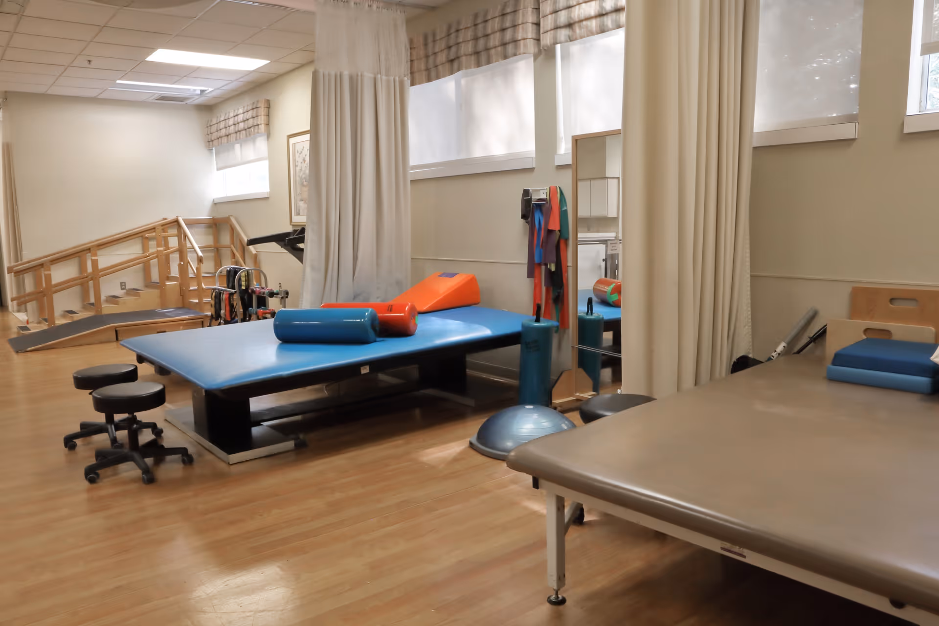 Physical therapy room with padded tables, therapy stools, exercise equipment including foam rollers, resistance bands, and a balance trainer. There is a wooden ramp and stairs in the background, with large windows covered by blinds letting in natural light.