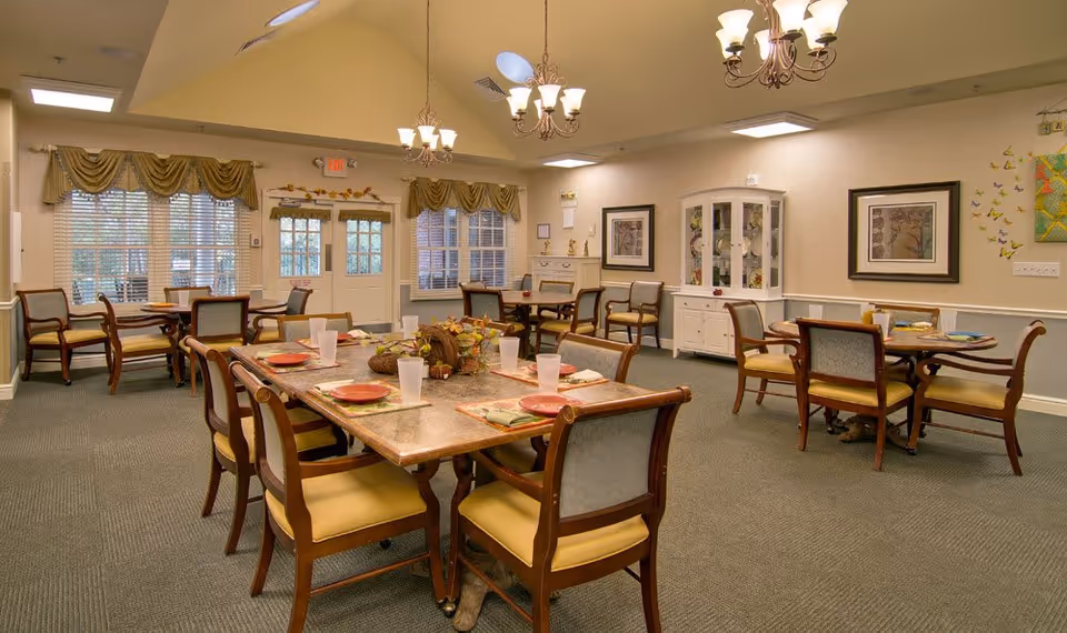 A well-lit senior dining room with multiple set tables and wooden chairs, chandeliers, and a display cabinet against the wall.