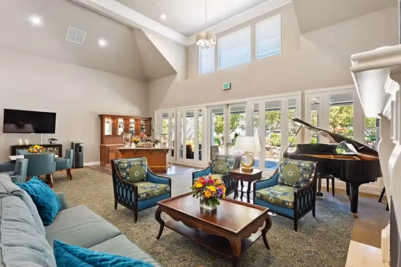 A spacious and well-lit living room area with high ceilings and large windows. The room features a light blue sofa with blue cushions, three patterned armchairs around a wooden coffee table with a flower arrangement, a grand piano, a side table with a lamp, and a wooden cabinet with glass doors. Outside greenery is visible through the glass doors.