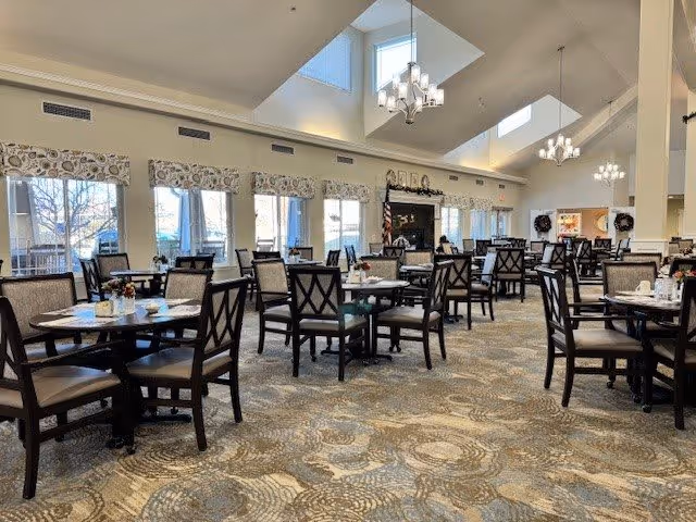 A spacious dining room with multiple round tables and cushioned chairs arranged neatly. Large windows with patterned valances allow natural light to fill the room. The ceiling features skylights and modern chandeliers. The carpet has a circular pattern design, and there is a fireplace with an American flag beside it in the background.