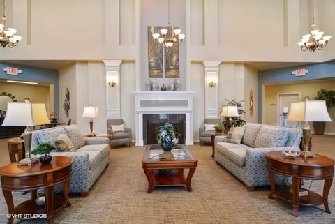 A spacious and well-lit living room area with two patterned sofas facing each other, two armchairs, a wooden coffee table with a floral centerpiece, and side tables with lamps. The room features a white fireplace with decorative items on the mantel, a large painting above it, and elegant chandeliers hanging from the high ceiling.