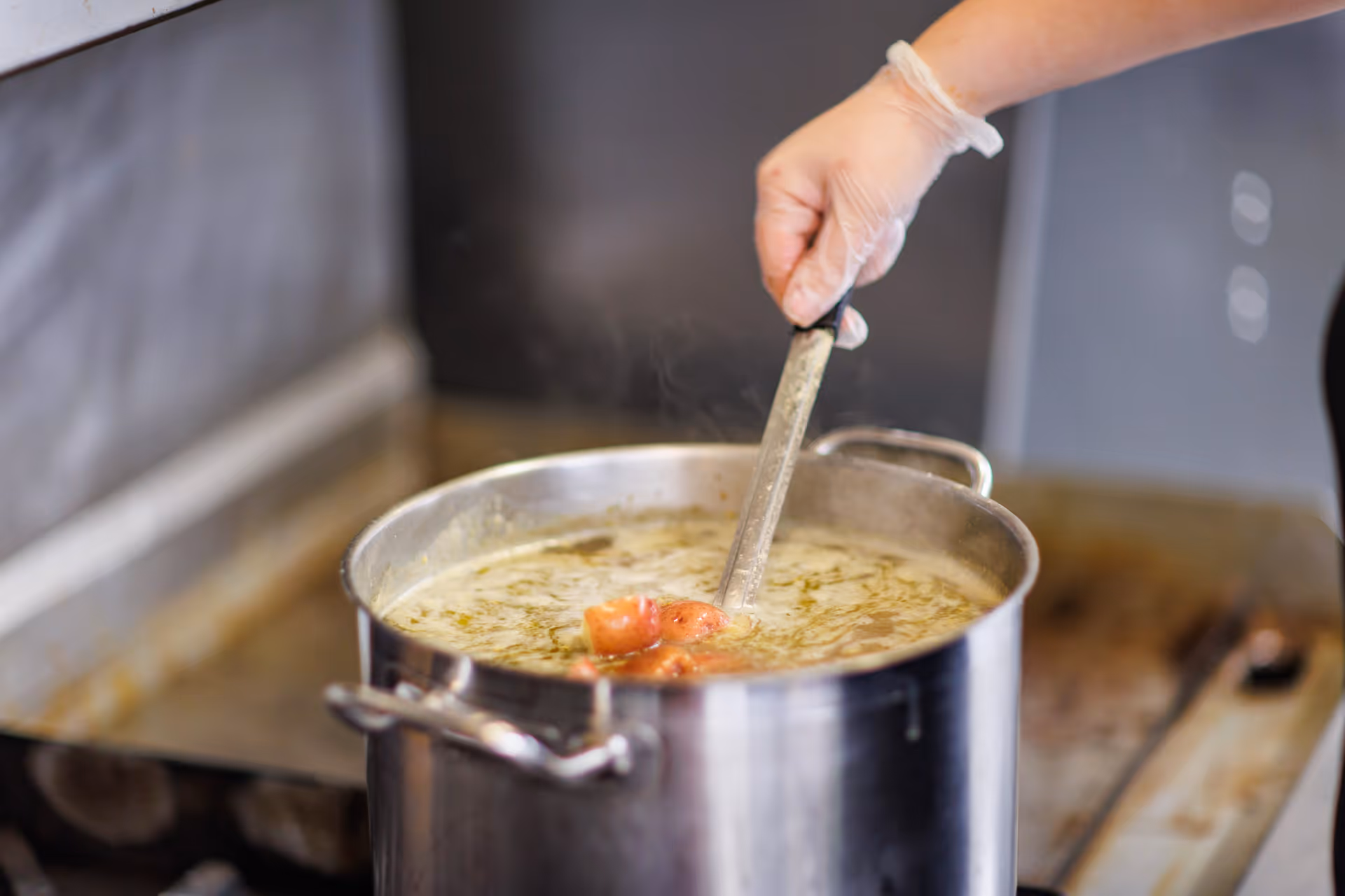 A hand wearing a disposable glove is stirring a large pot of soup or stew with a ladle in a kitchen setting.