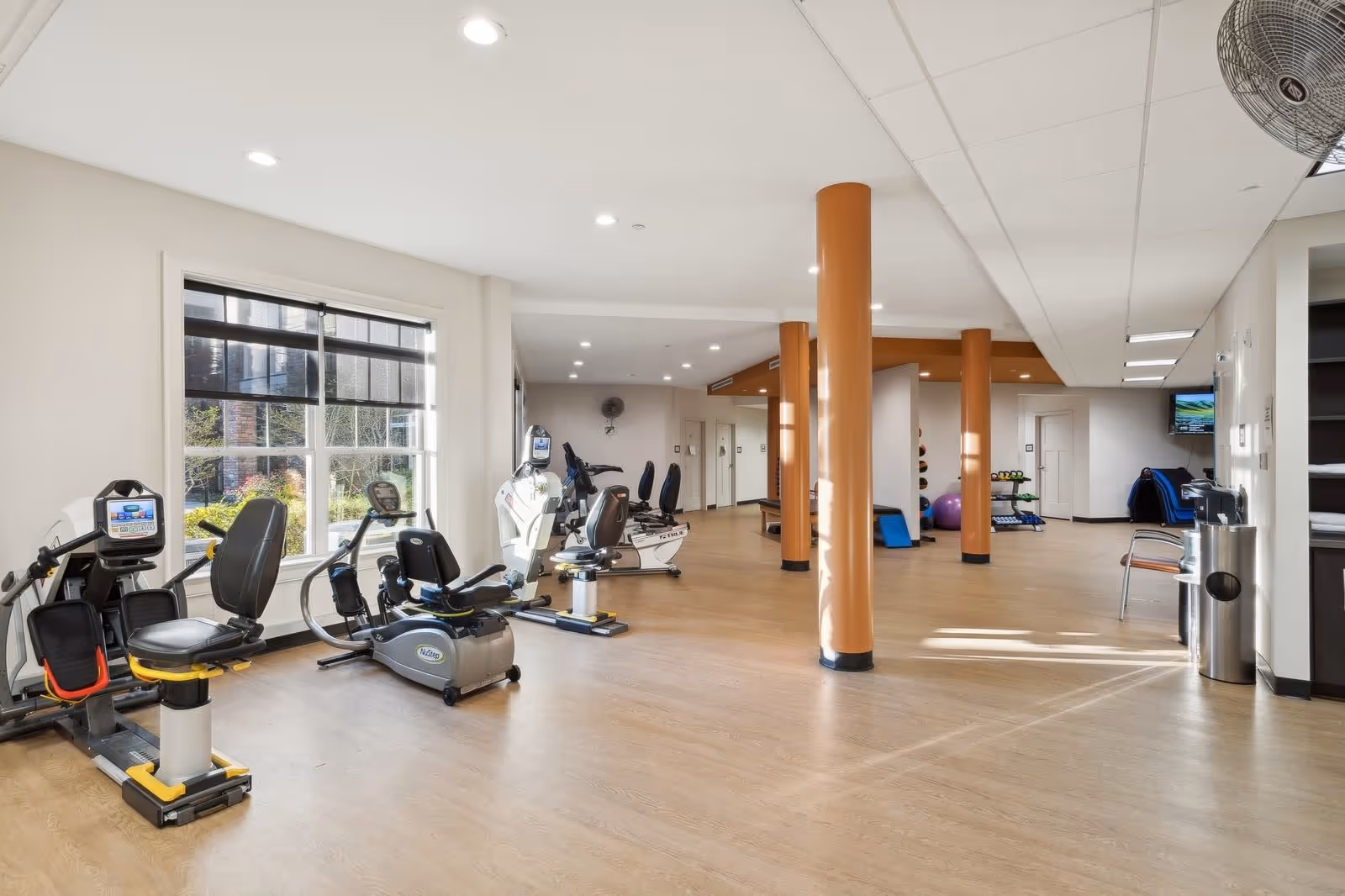 A spacious fitness room with exercise equipment including stationary bikes and rowing machines, large windows letting in natural light, wooden flooring, and orange support columns. There are also exercise balls and weights visible in the background.