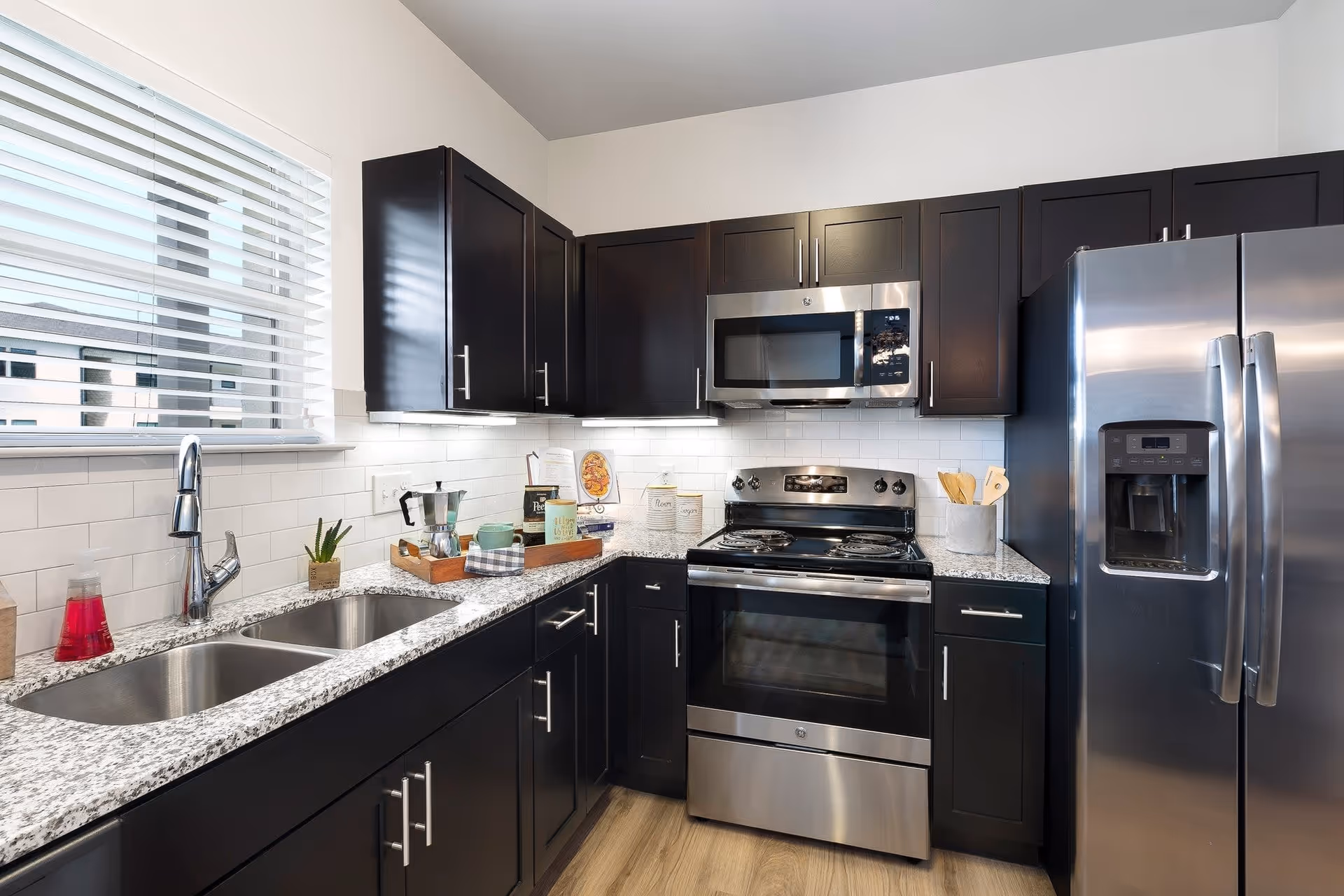 Modern kitchen with dark wood cabinets, granite countertops, stainless steel appliances including a refrigerator, stove, and microwave. There is a double sink under a window with blinds, and various kitchen items like a coffee maker, mugs, and utensils on the counter.
