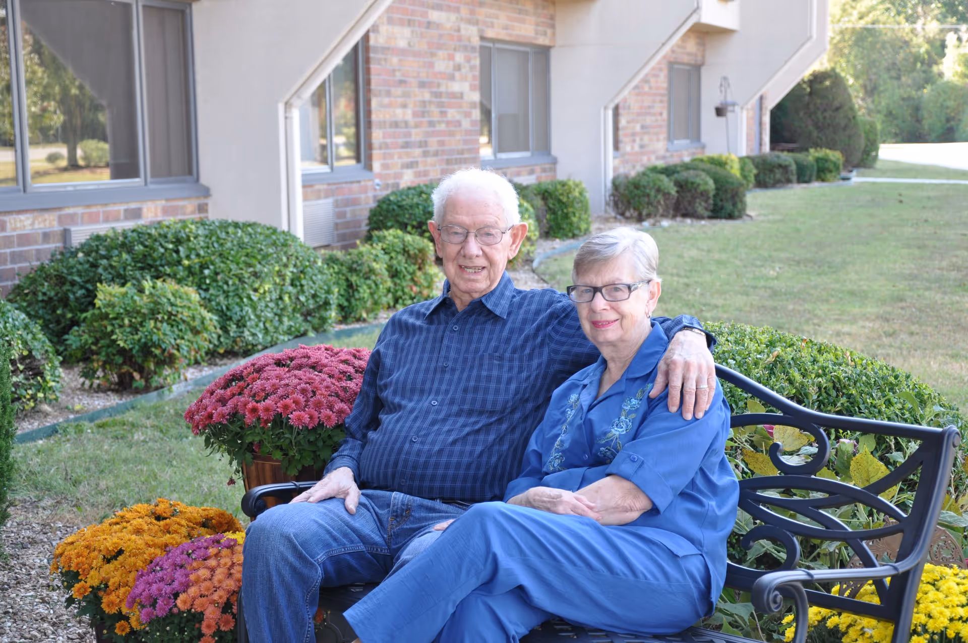 An elderly couple sitting on a black metal bench outside a brick building with neatly trimmed bushes and colorful flower pots around them. The man has white hair and glasses, wearing a blue checkered shirt and jeans, with his arm around the woman who is wearing glasses and a blue outfit. They are smiling and appear relaxed in a garden-like setting.