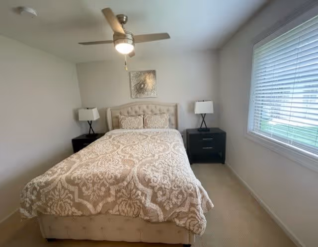 A bedroom with a beige patterned bedspread on a bed with a tufted headboard. There are two black nightstands on either side of the bed, each with a lamp. A ceiling fan with a light is above the bed, and a window with blinds is on the right wall. A decorative piece of art hangs above the headboard.