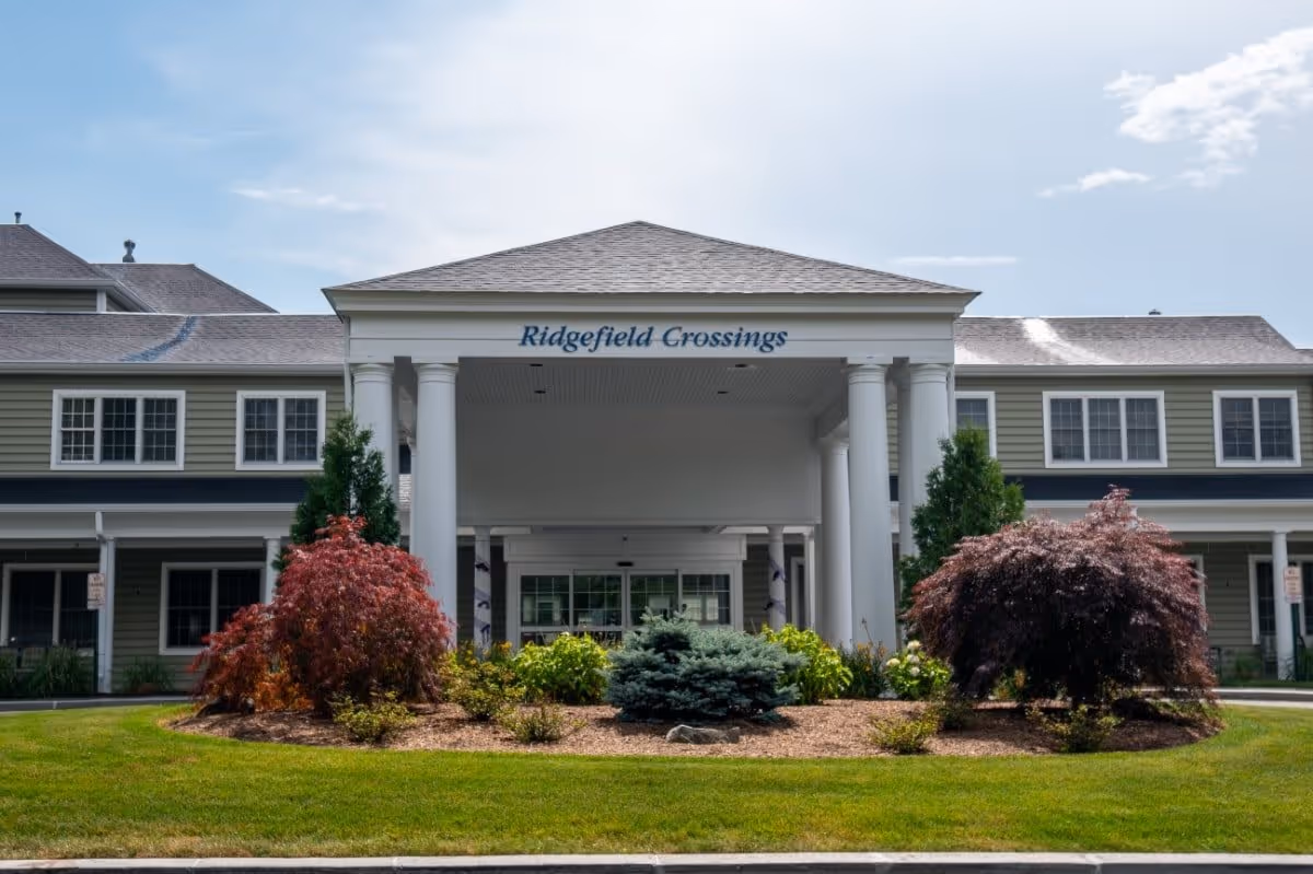 Front exterior view of Ridgefield Crossings building with a covered entrance supported by white columns, surrounded by landscaped bushes and green grass under a partly cloudy sky.