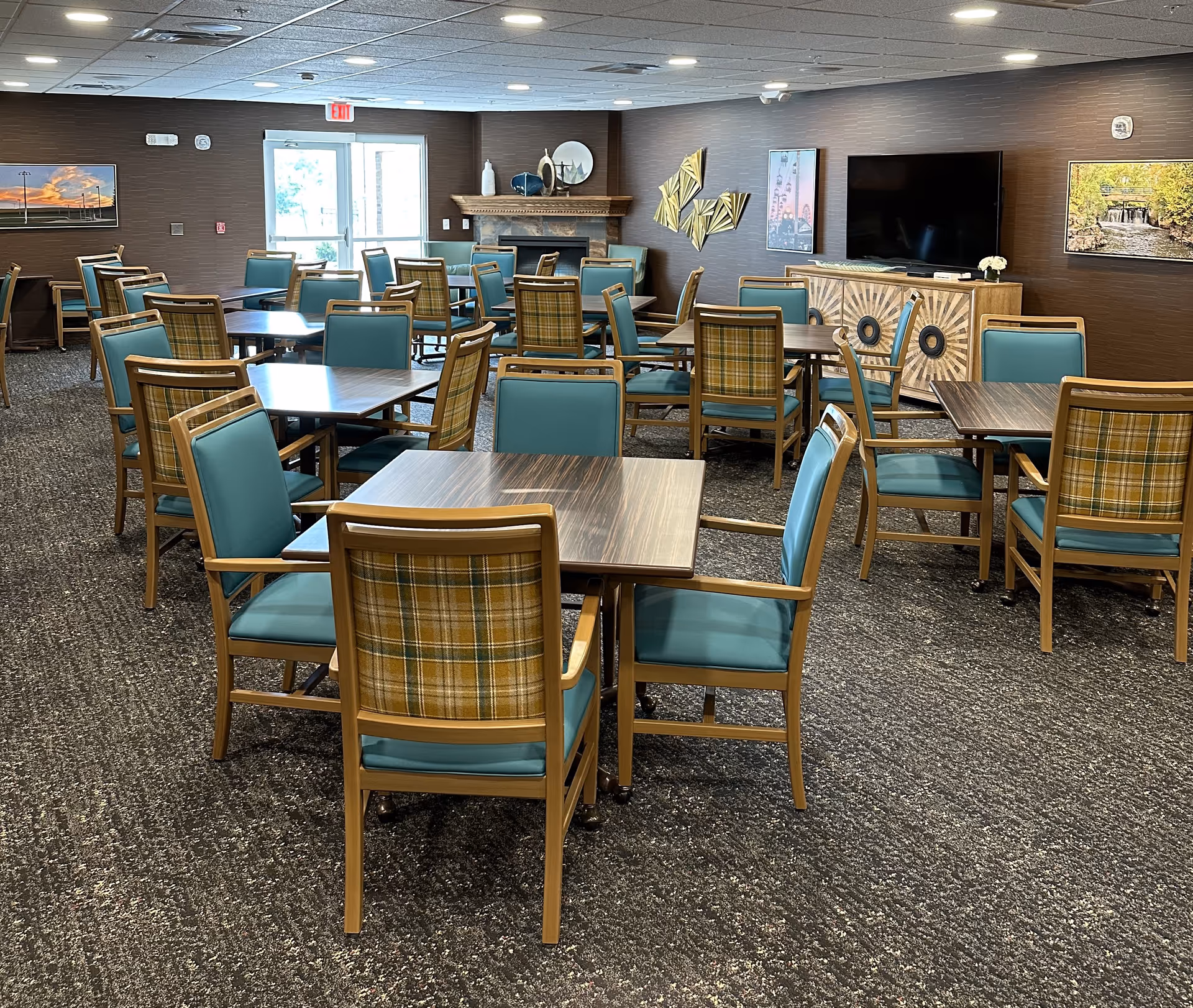 Spacious dining/activity room with multiple square tables and teal-upholstered chairs arranged on carpet facing a TV and fireplace.