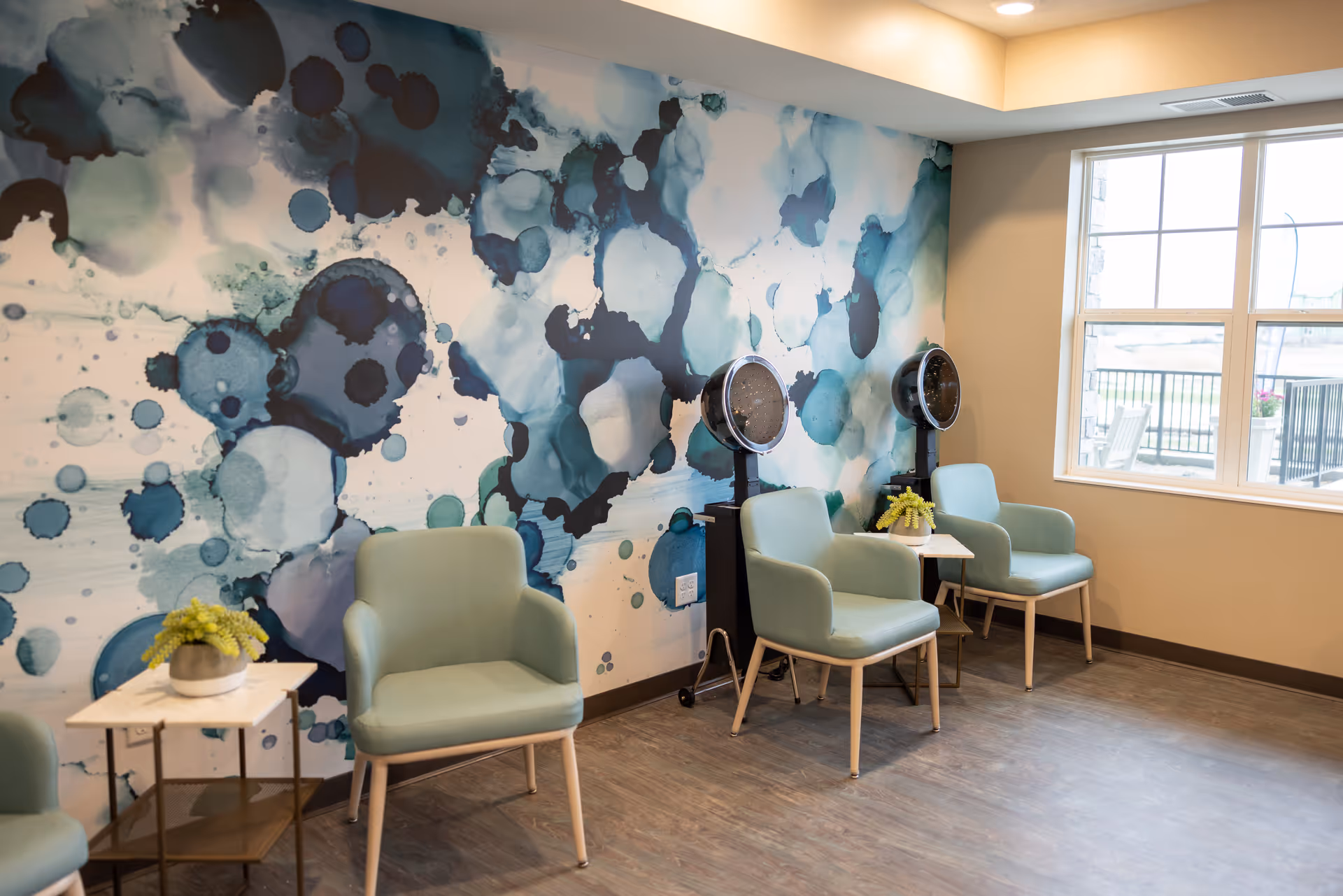 A bright room with three light green armchairs arranged along a wall with a blue and teal abstract watercolor mural. Two vintage hair dryers are positioned behind two of the chairs. Small side tables with potted plants are placed between the chairs. A large window lets in natural light, showing an outdoor patio area.