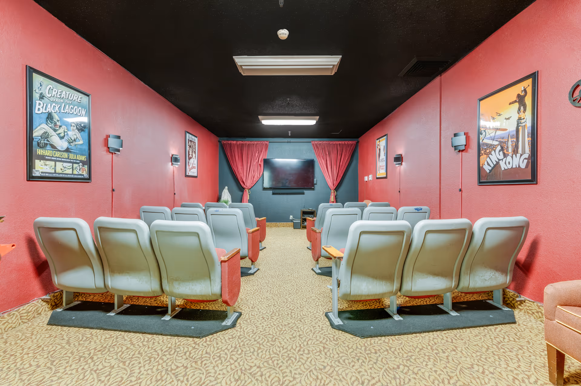 A small movie theater room with two rows of gray theater seats facing a wall-mounted flat screen TV. The walls are painted red with movie posters including 'Creature from the Black Lagoon' and 'King Kong'. The floor is carpeted with a patterned design and there are red curtains on either side of the TV.