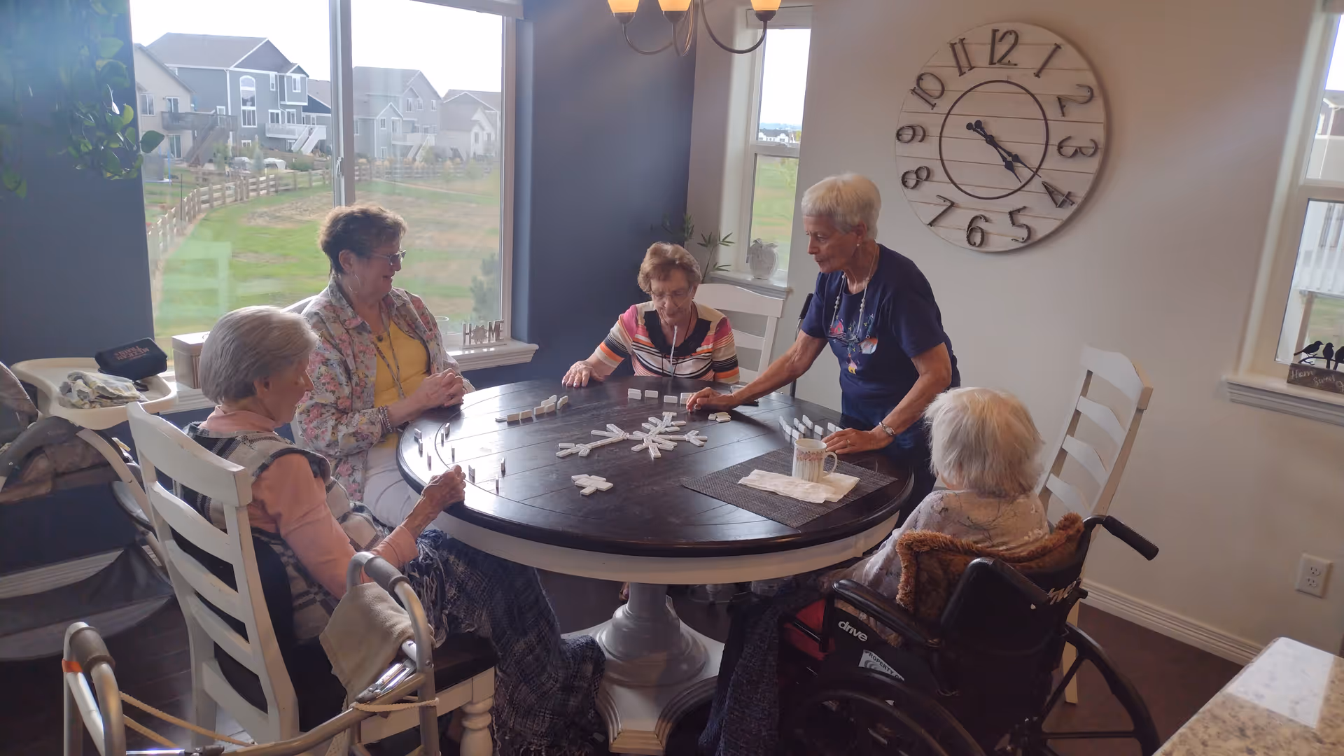 Five elderly women sitting around a round wooden table playing a game of dominoes in a well-lit room with large windows showing a view of houses and green lawns outside. One woman is in a wheelchair, and there is a large wall clock behind them.