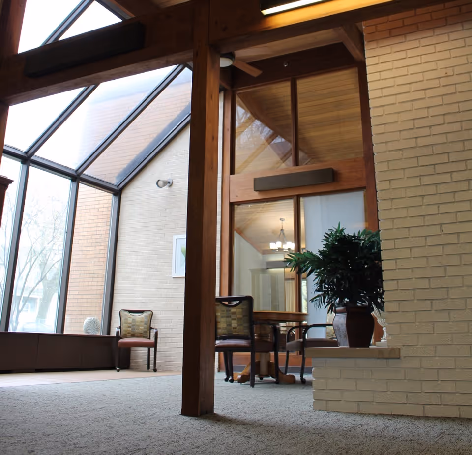 Interior view of a senior living facility with large angled windows letting in natural light, a carpeted floor, wooden beams, a potted plant on a ledge, and several chairs around a small table in the background.