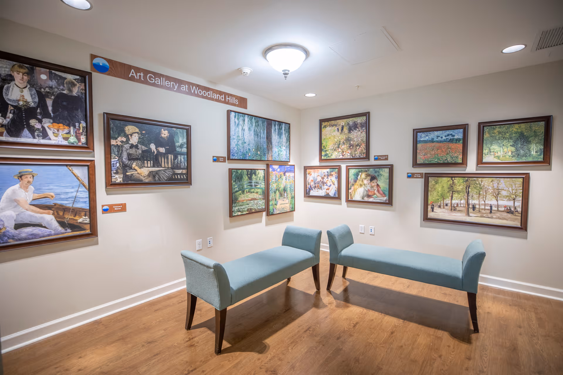 Interior view of an art gallery at Woodland Hills featuring multiple framed paintings on cream-colored walls. Two light blue upholstered benches with wooden legs are placed on a wooden floor beneath a ceiling light fixture.