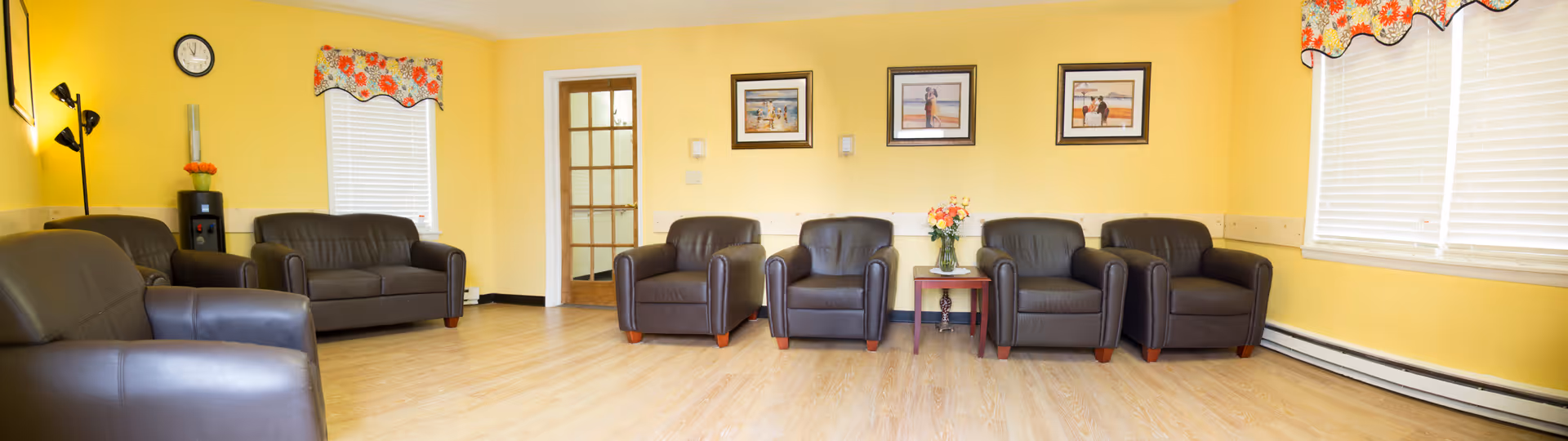 A spacious living room with yellow walls and light wood flooring, furnished with multiple dark brown leather armchairs and a matching loveseat. The room has two windows with white blinds and floral valances, a small table with a flower vase, three framed pictures on the wall, a clock, and a floor lamp in the corner.