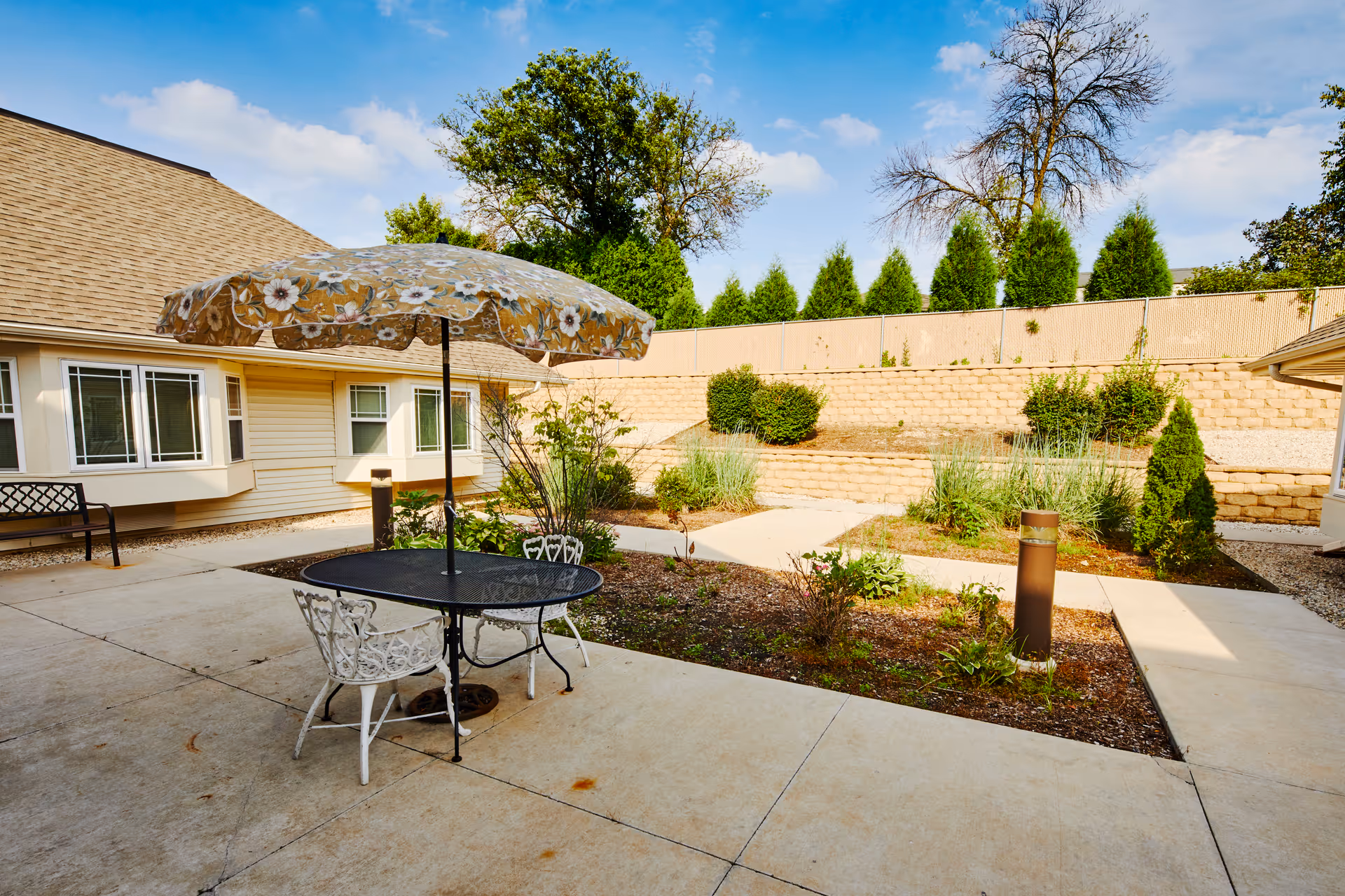 Outdoor patio area with a round black metal table and two white chairs under a floral-patterned umbrella. The patio is surrounded by a garden with various plants and shrubs, a beige building with windows, and a stone retaining wall with trees and bushes in the background under a blue sky with some clouds.