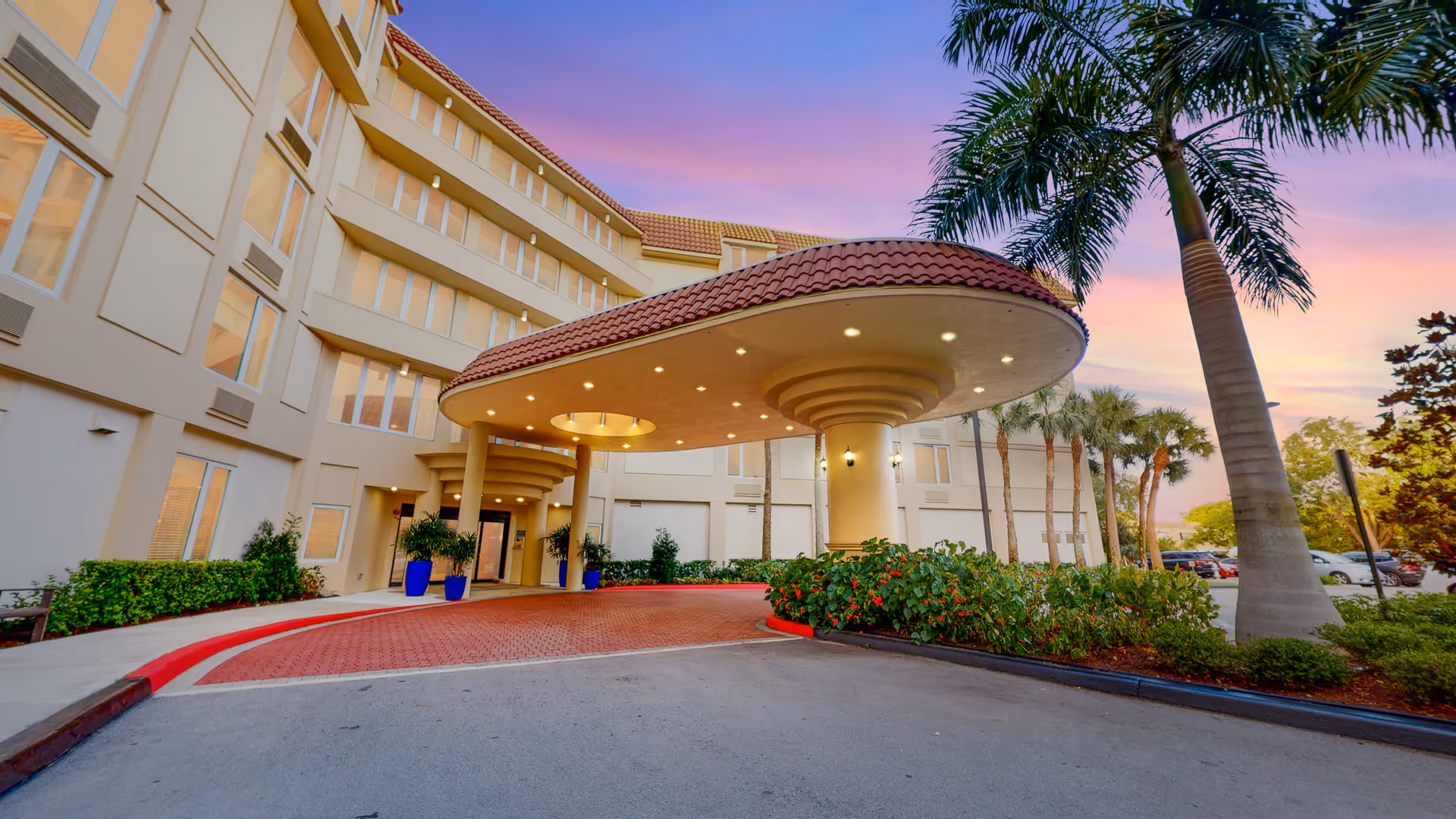 Exterior view of The Atrium at Boca Raton senior living facility entrance with a covered circular driveway, palm trees, and a colorful sunset sky in the background.