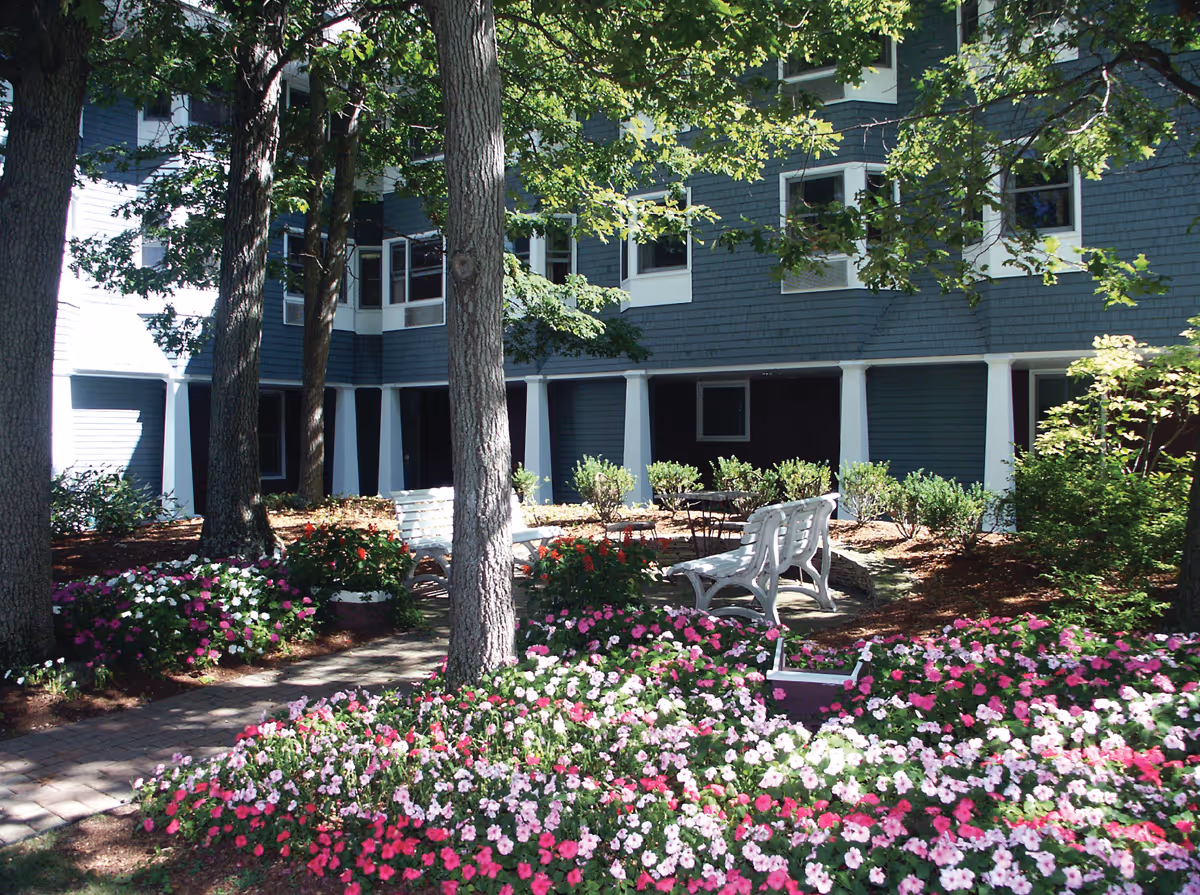 Outdoor garden area with colorful flowers, trees, white benches, and a building with blue siding and white pillars in the background.