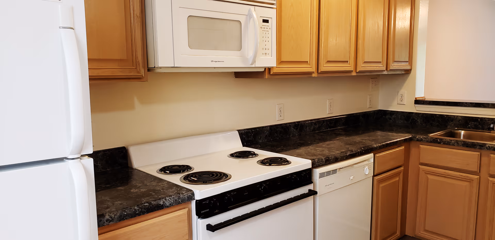 Kitchen with white refrigerator, microwave, electric stove, and dishwasher, wood cabinets and dark marble-look countertops.