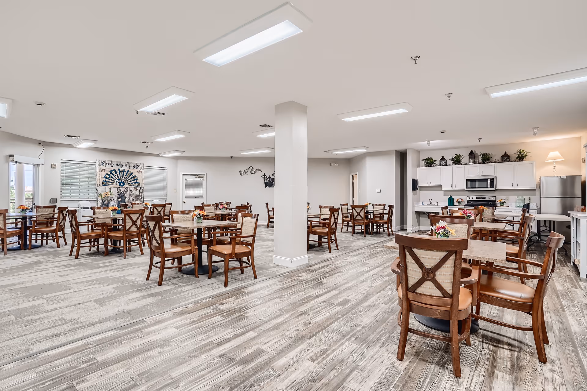 A spacious dining room in a senior living facility with multiple wooden tables and chairs arranged neatly. The room has light-colored walls, large windows with blinds, and a kitchen area with white cabinets, a microwave, and a refrigerator. There are decorative plants and flowers on the tables and shelves, and the floor has a wood-like finish.