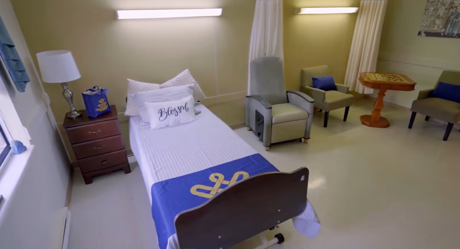 A clean and simple room with a single hospital-style bed covered with white linens and a blue blanket at the foot. The bed has pillows, including one with the word 'Blessed' embroidered on it. Next to the bed is a wooden nightstand with a lamp and a blue bag. The room also features two armchairs with blue cushions and a wooden table with a checkerboard game on it. The walls are light-colored, and there are two horizontal wall lights above the bed and chairs. A window with blue curtains is visible on the left side.